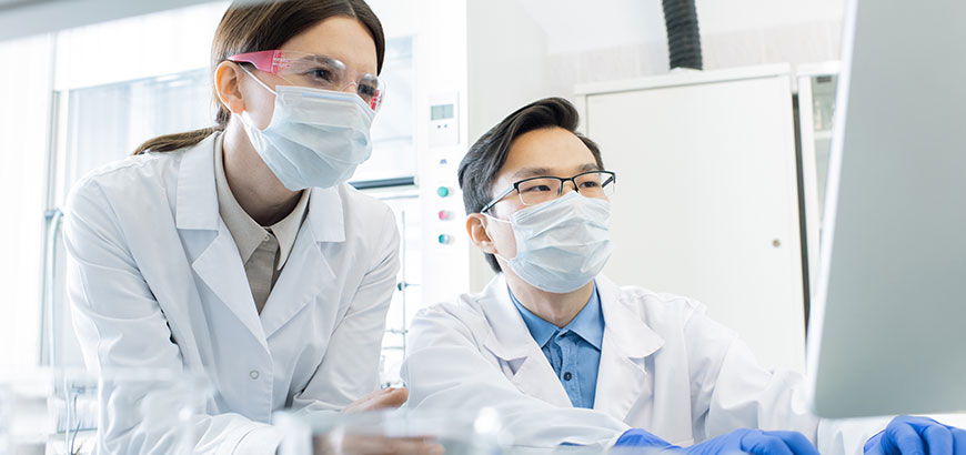 Two pharmacists wearing masks reviewing data on a computer monitor