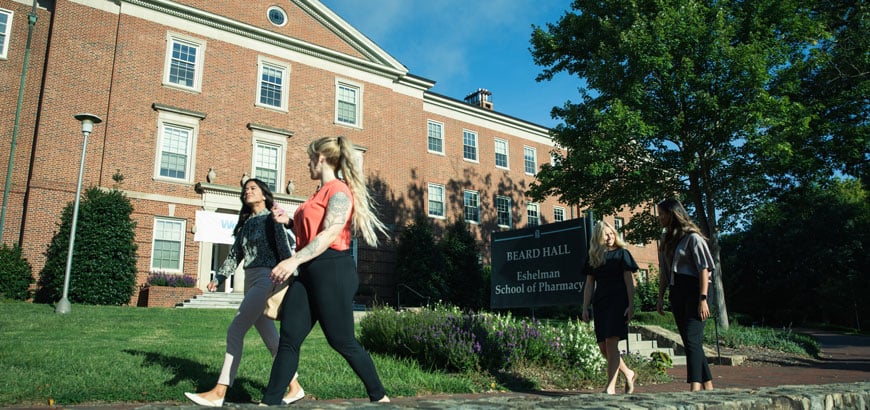 Two women walk down a sidewalk past a pharmacy school.