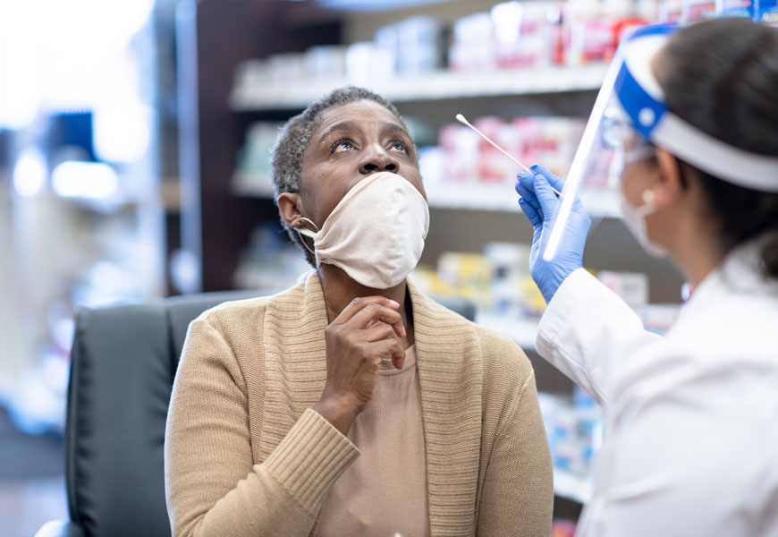 A pharmacist in full PPE puts a long q-tip up a patient's nose to test for COVID 19